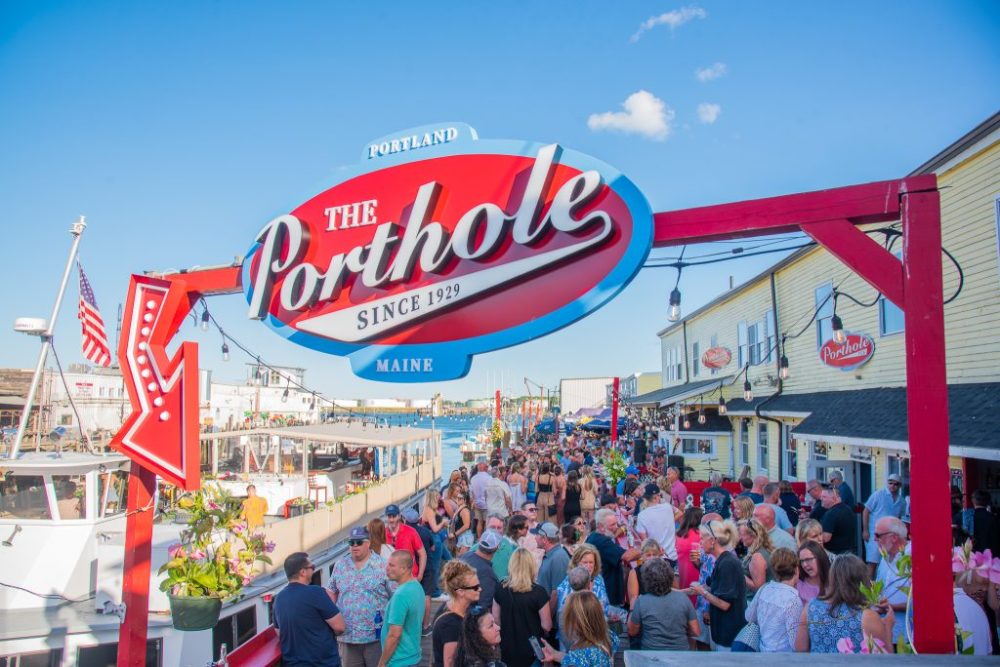 Crowd gathers under 'The Porthole' sign in Portland, Maine, with boats and buildings visible in background.