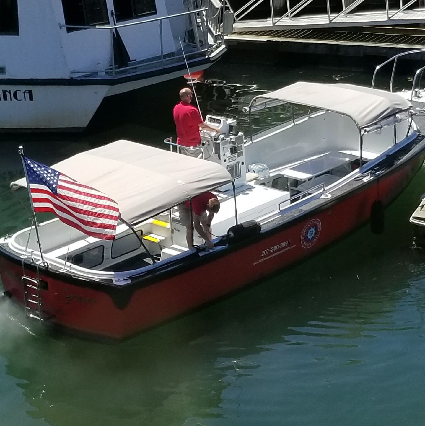 Red boat with US flag and two people onboard near dock.