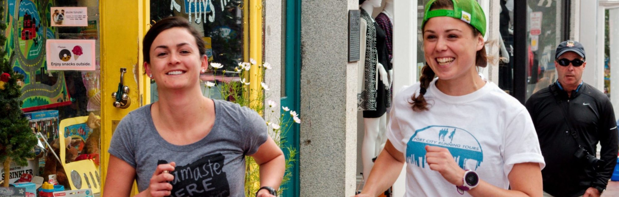 Two women running and smiling near shop; a man walking behind them.