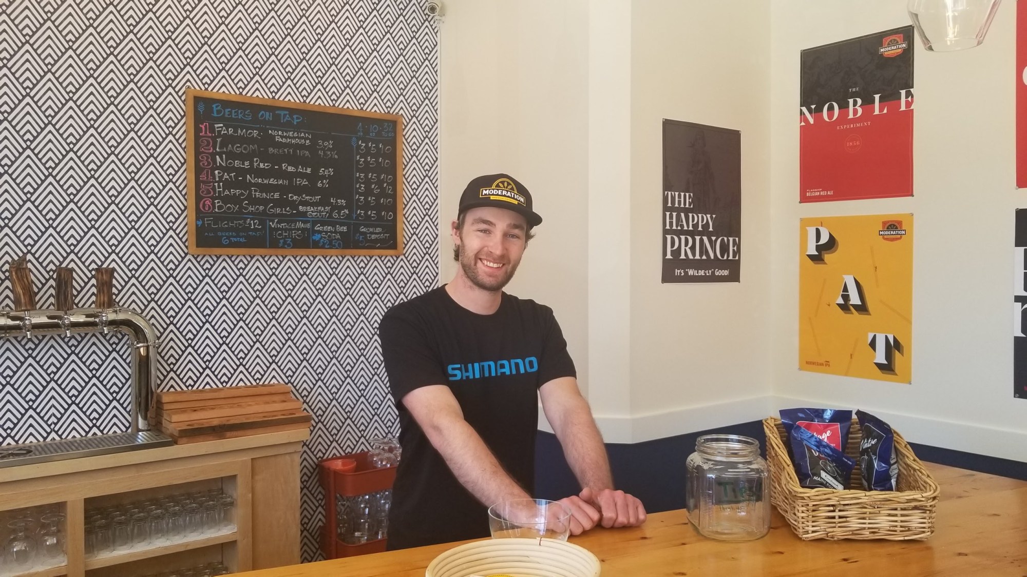Man smiling behind a bar with a beer tap and menu on the wall.