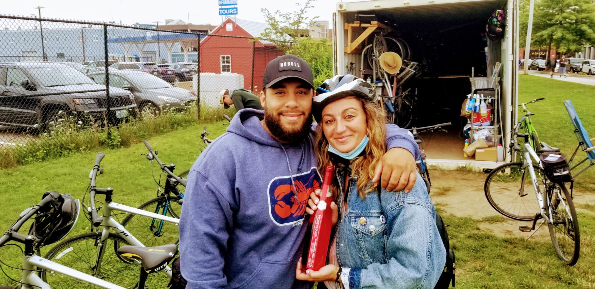Two people in front of open container with bikes; one holding a red item, wearing a helmet and denim jacket.