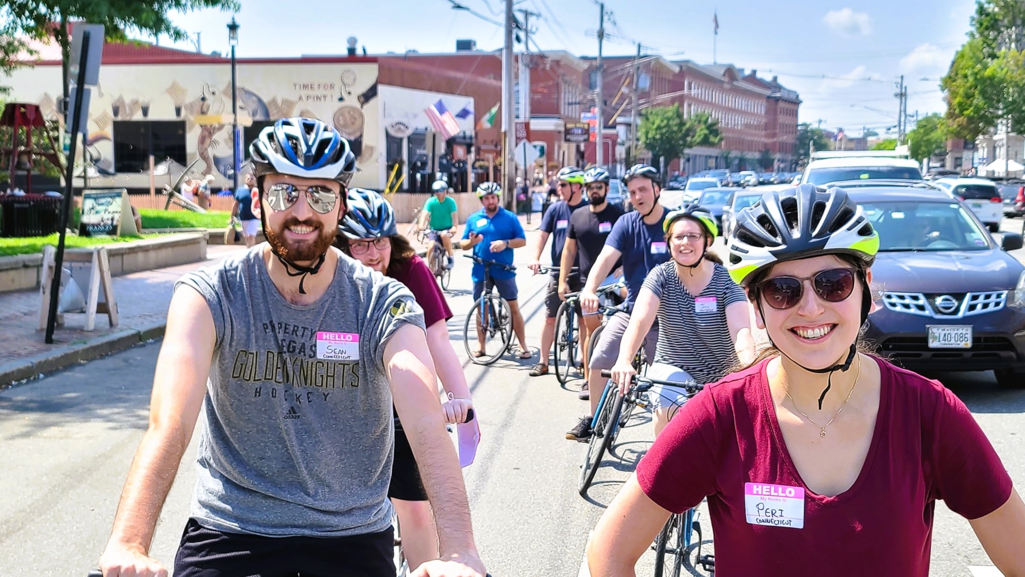 Group of cyclists wearing helmets and name tags biking on a sunny day in an urban area.
