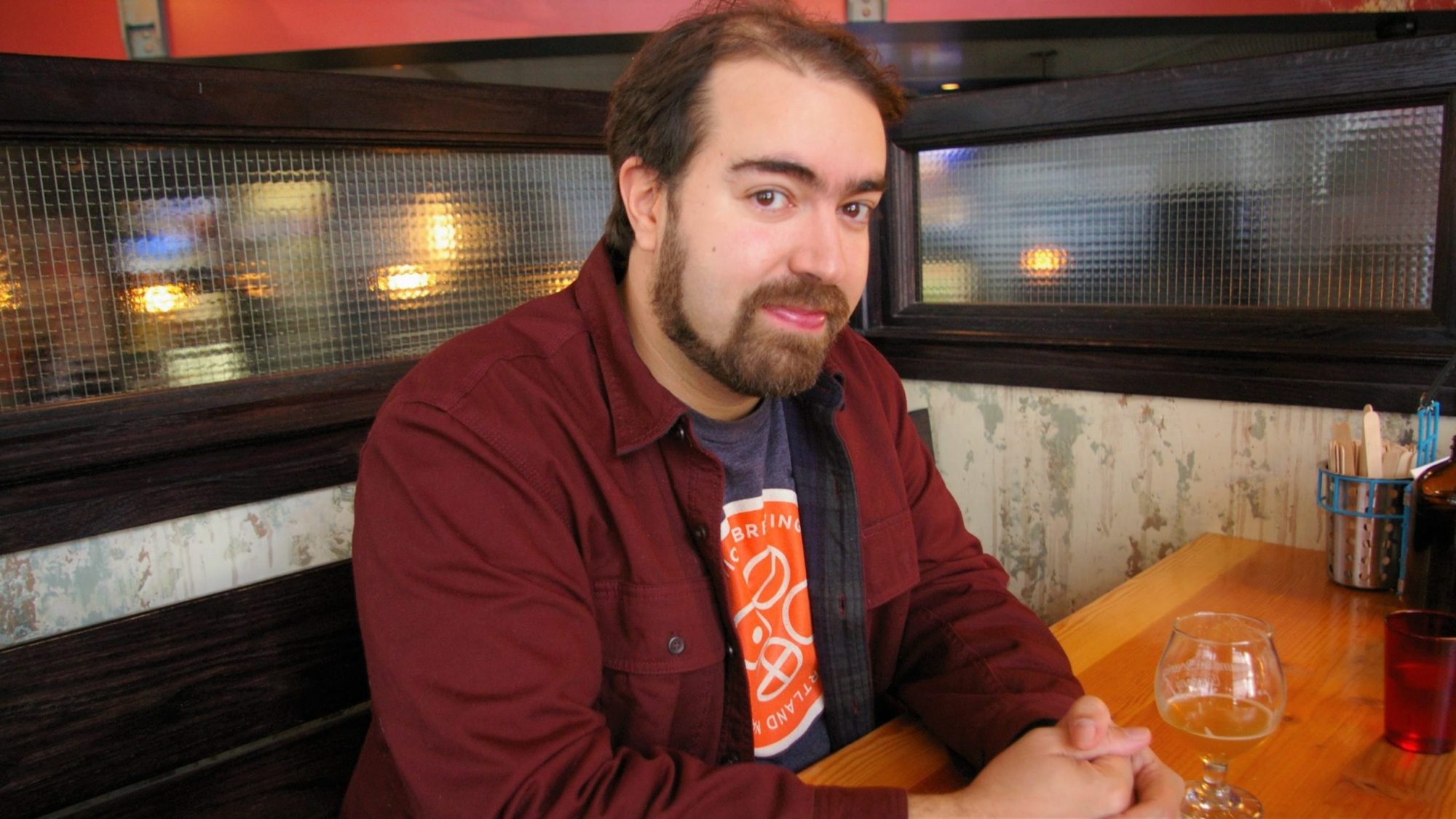 Bearded man in a burgundy jacket seated at a wooden table in a cozy restaurant.