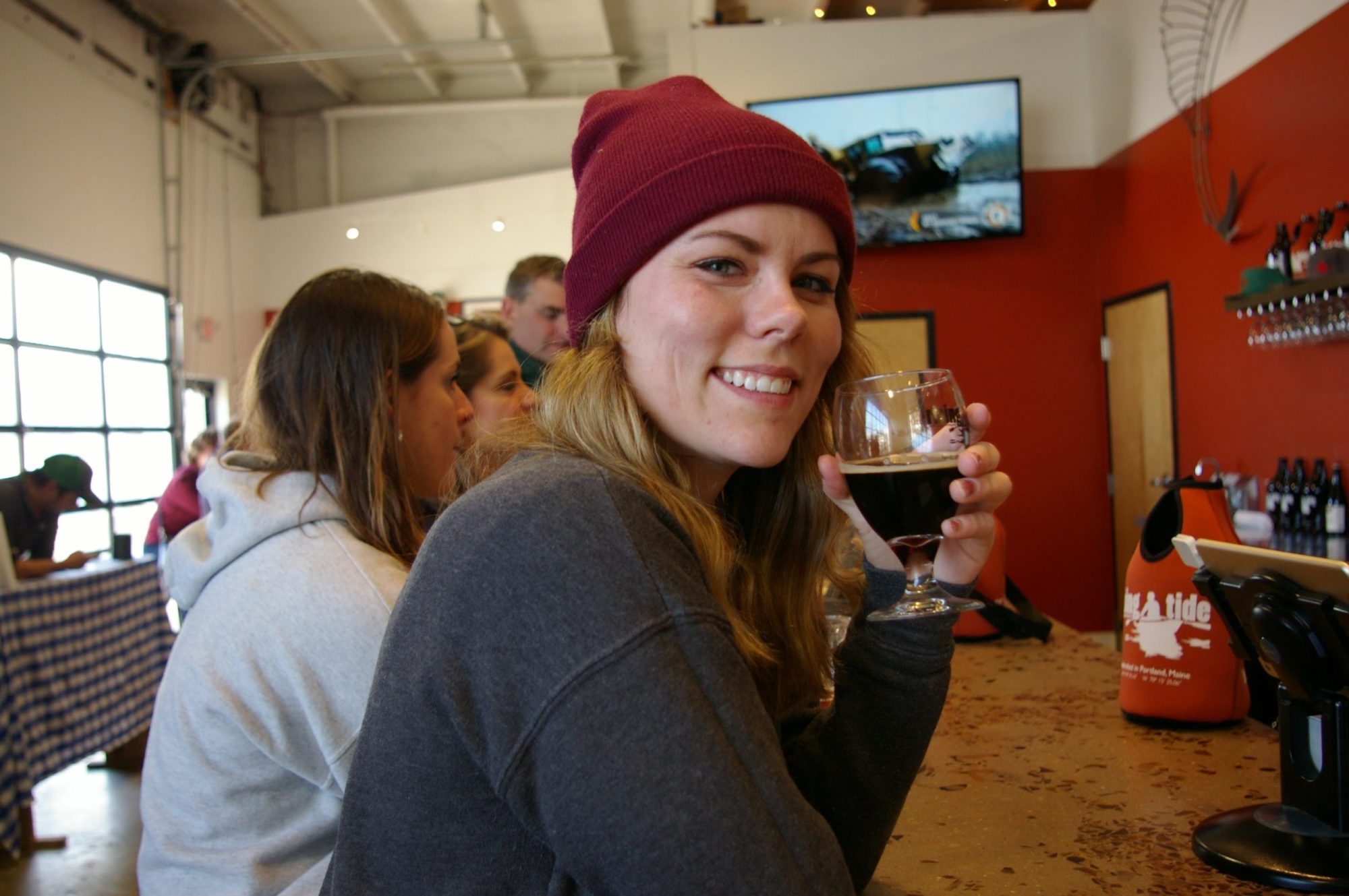Woman in red beanie smiles, holding a glass of beer at a bar counter.