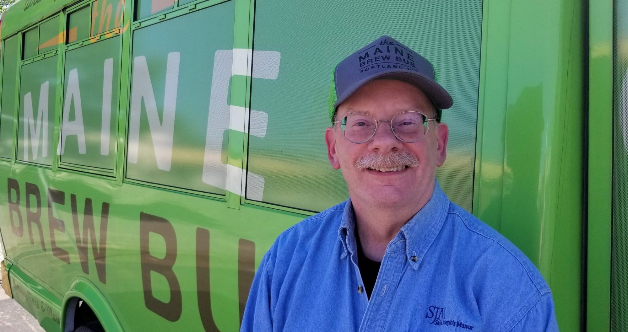 Man in glasses and cap smiling beside green bus with 'Maine Brew Bus' logo.