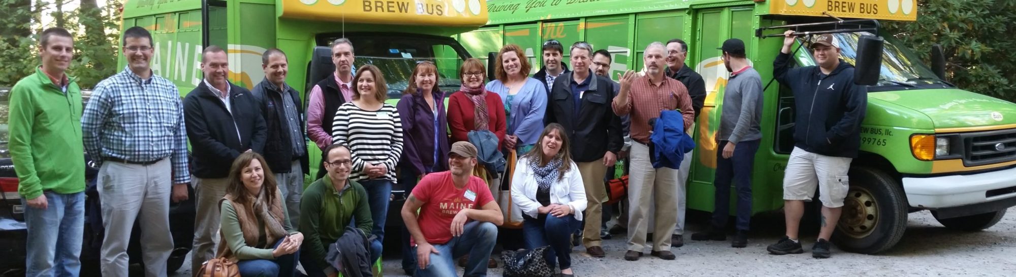 Group of people posing in front of a green 'brew bus' in a wooded area.
