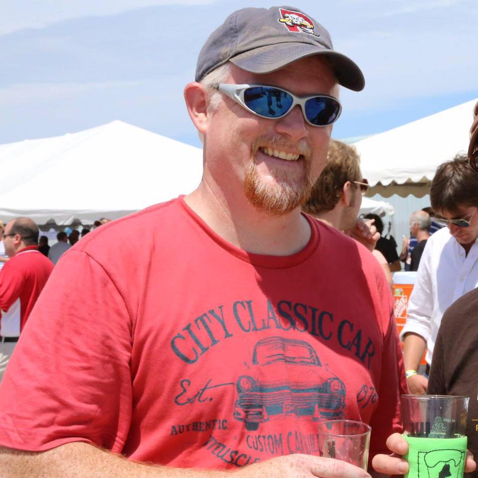 Man in a red shirt and cap smiles holding a drink outdoors.