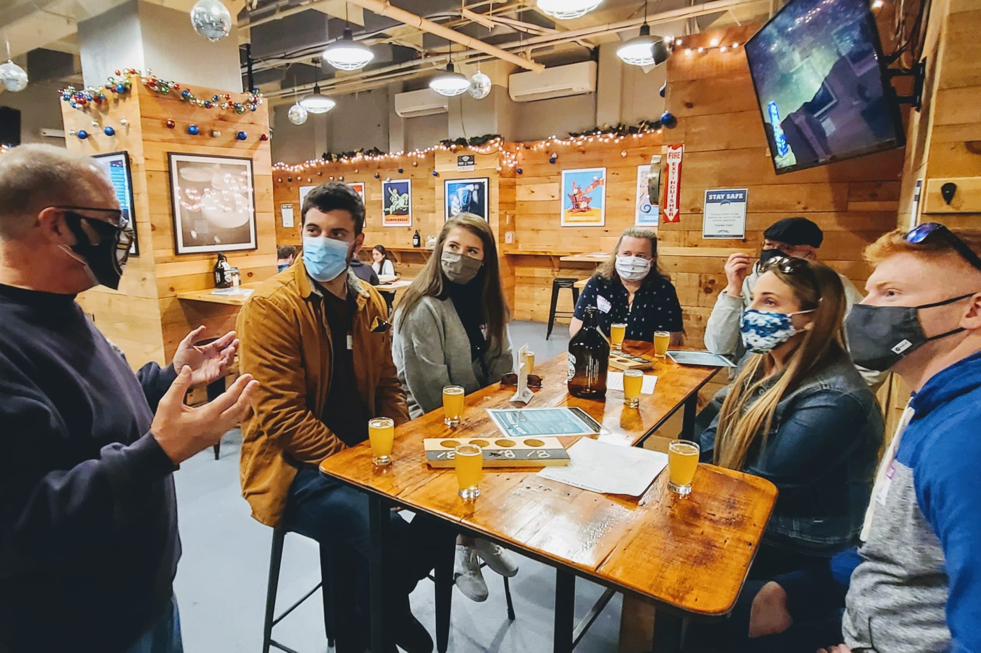 People in masks sit around a wooden table with drinks in a festive, warmly lit room.