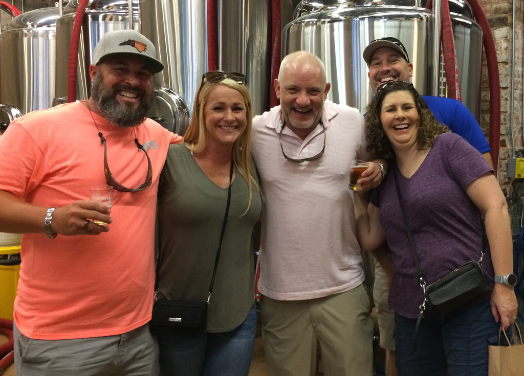 Group of five smiling people posing in front of brewery tanks.