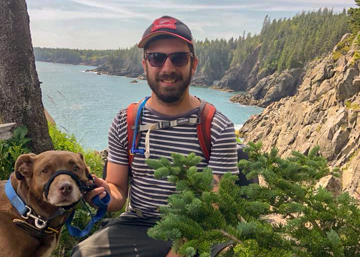 Man with a cap and sunglasses sitting with a dog by a coastal cliff.