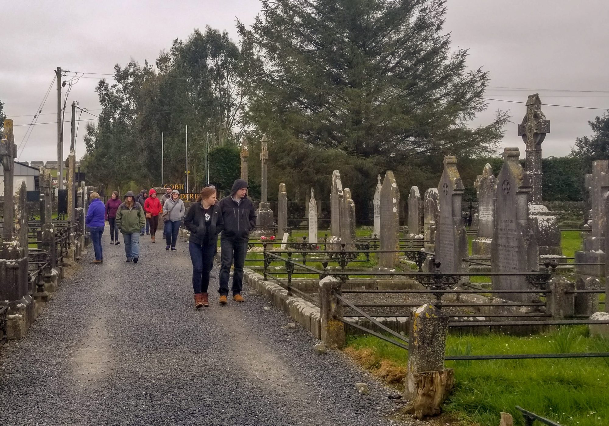 People walking on a gravel path in a cemetery with headstones and trees.