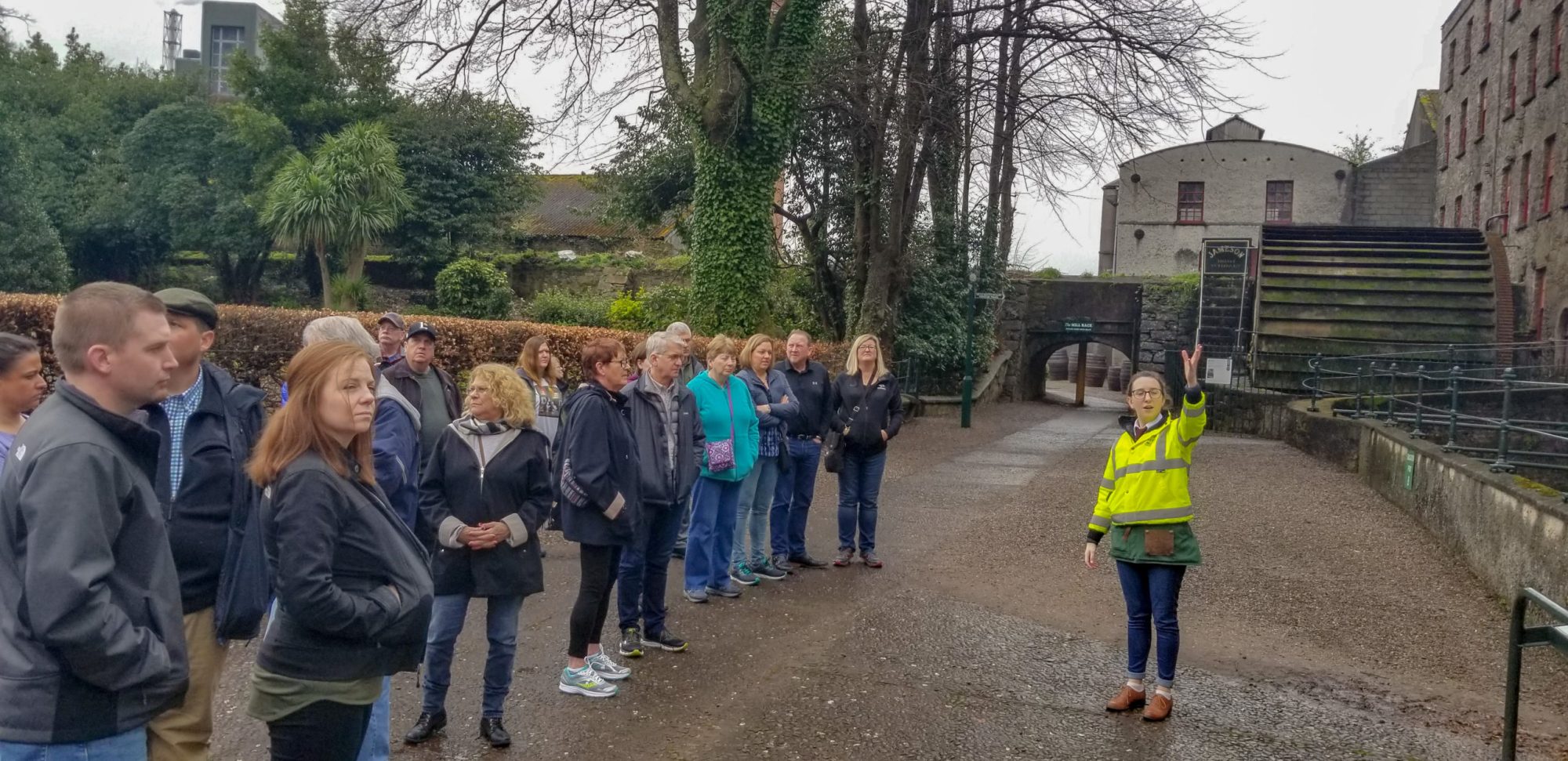 Tour guide in yellow jacket leads a group of people near a historic site with trees and a stone building.