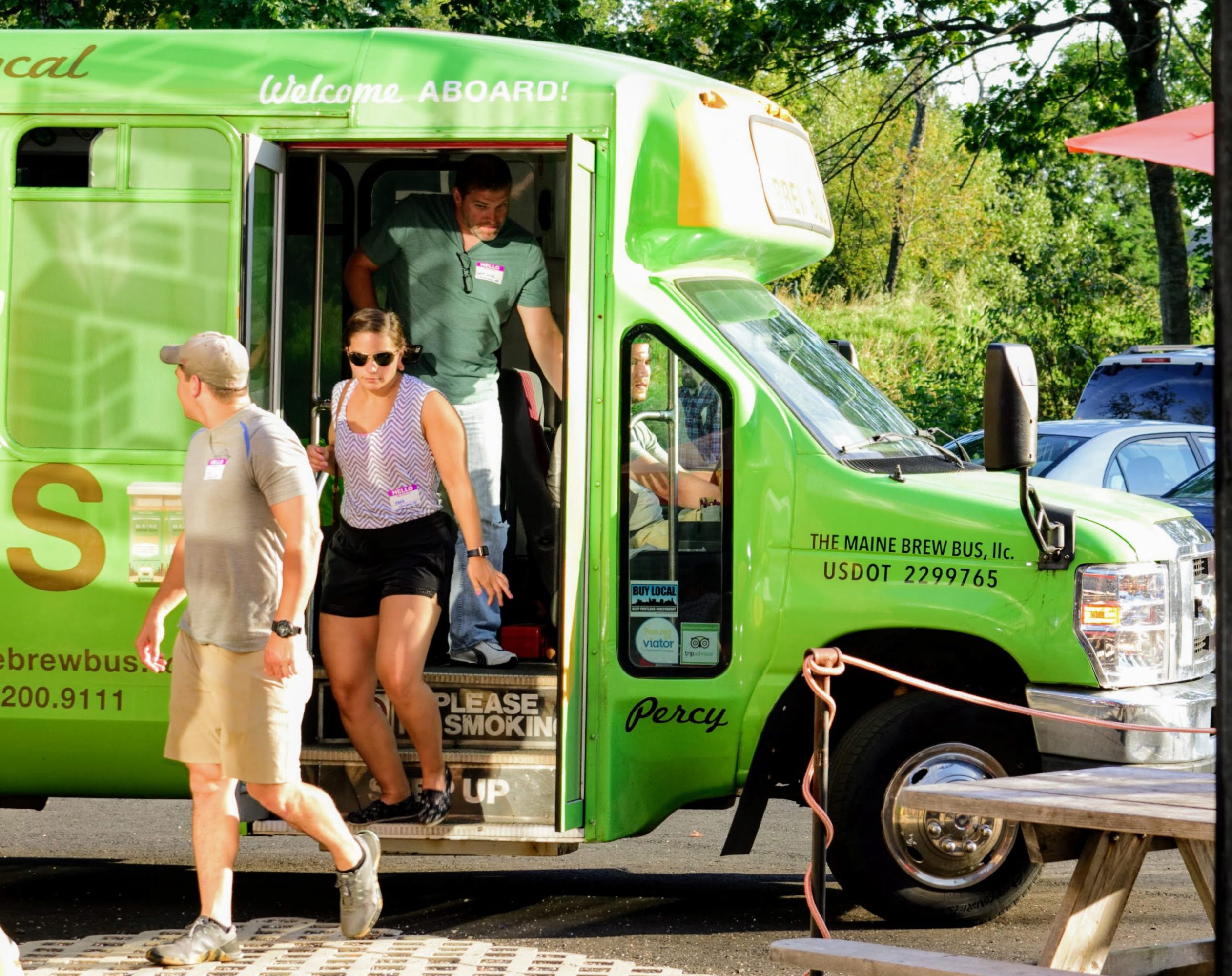 People exiting a green tour bus labeled 'The Maine Brew Bus' in a sunny parking area.