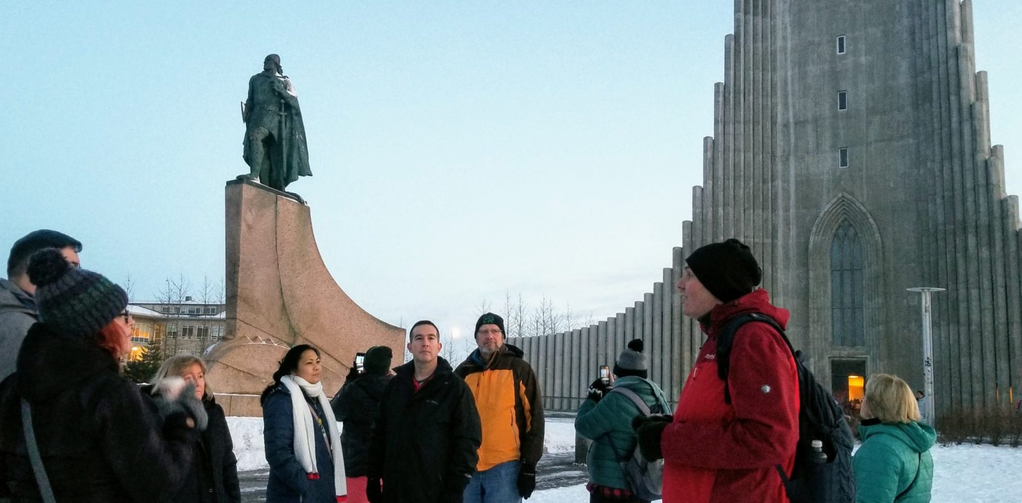 Group of people sightseeing near a tall building and statue on a snowy day.
