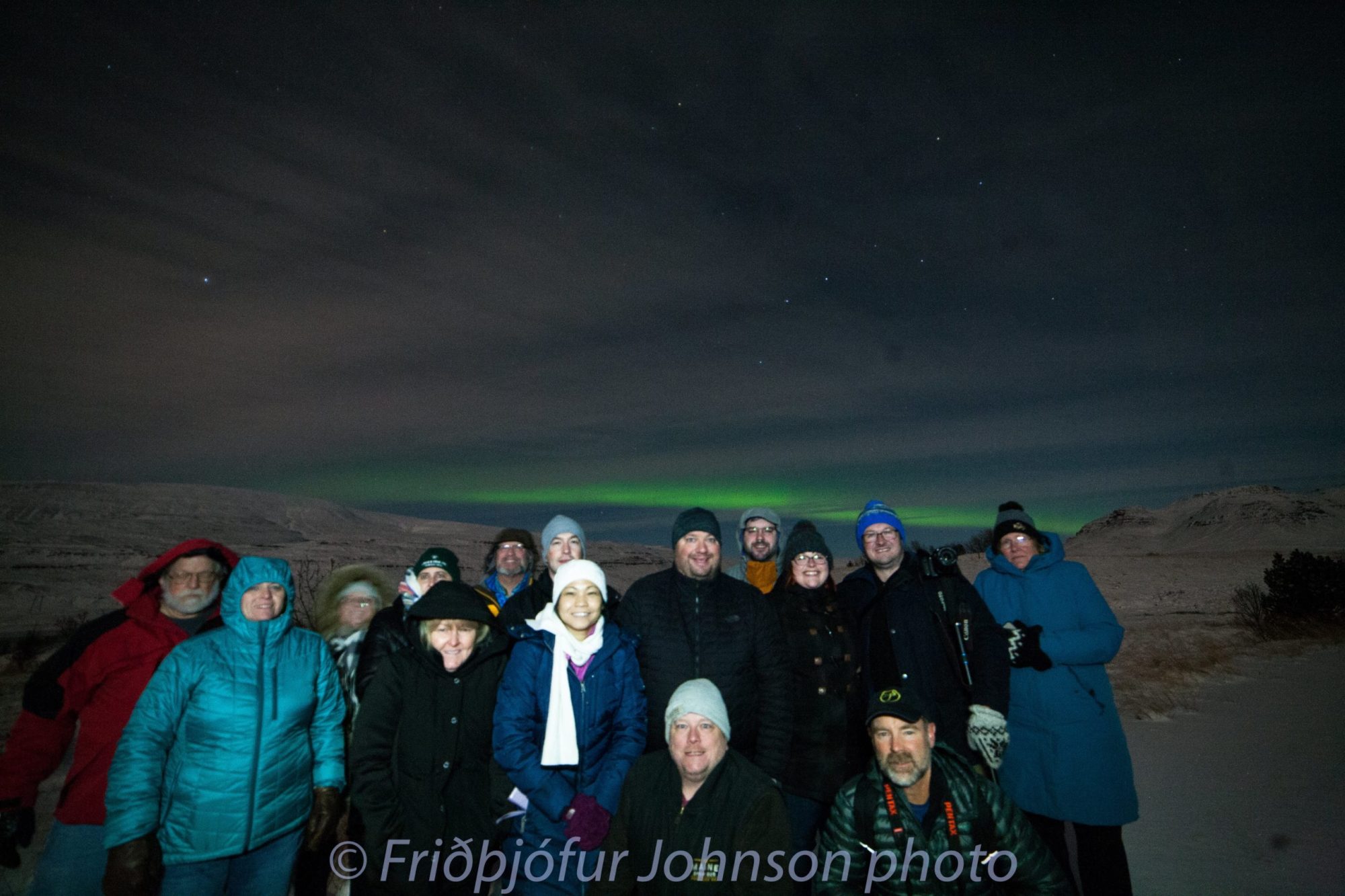 Group of people in winter clothing under Northern Lights in a snowy landscape.
