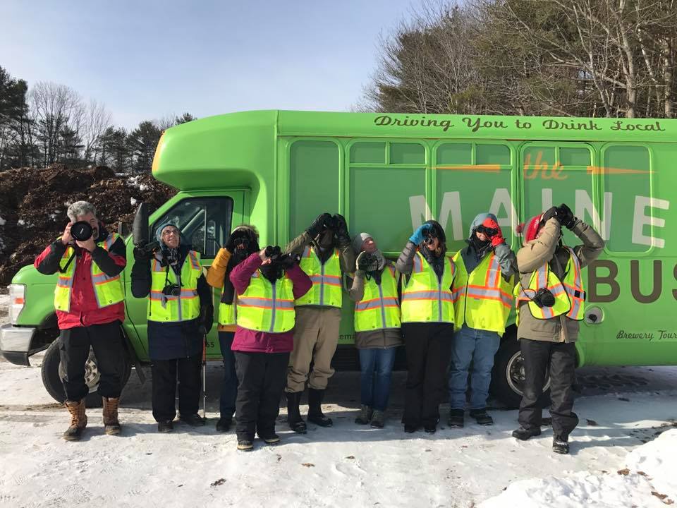 Group in bright vests posing with cameras in front of a green tour bus on a snowy day.
