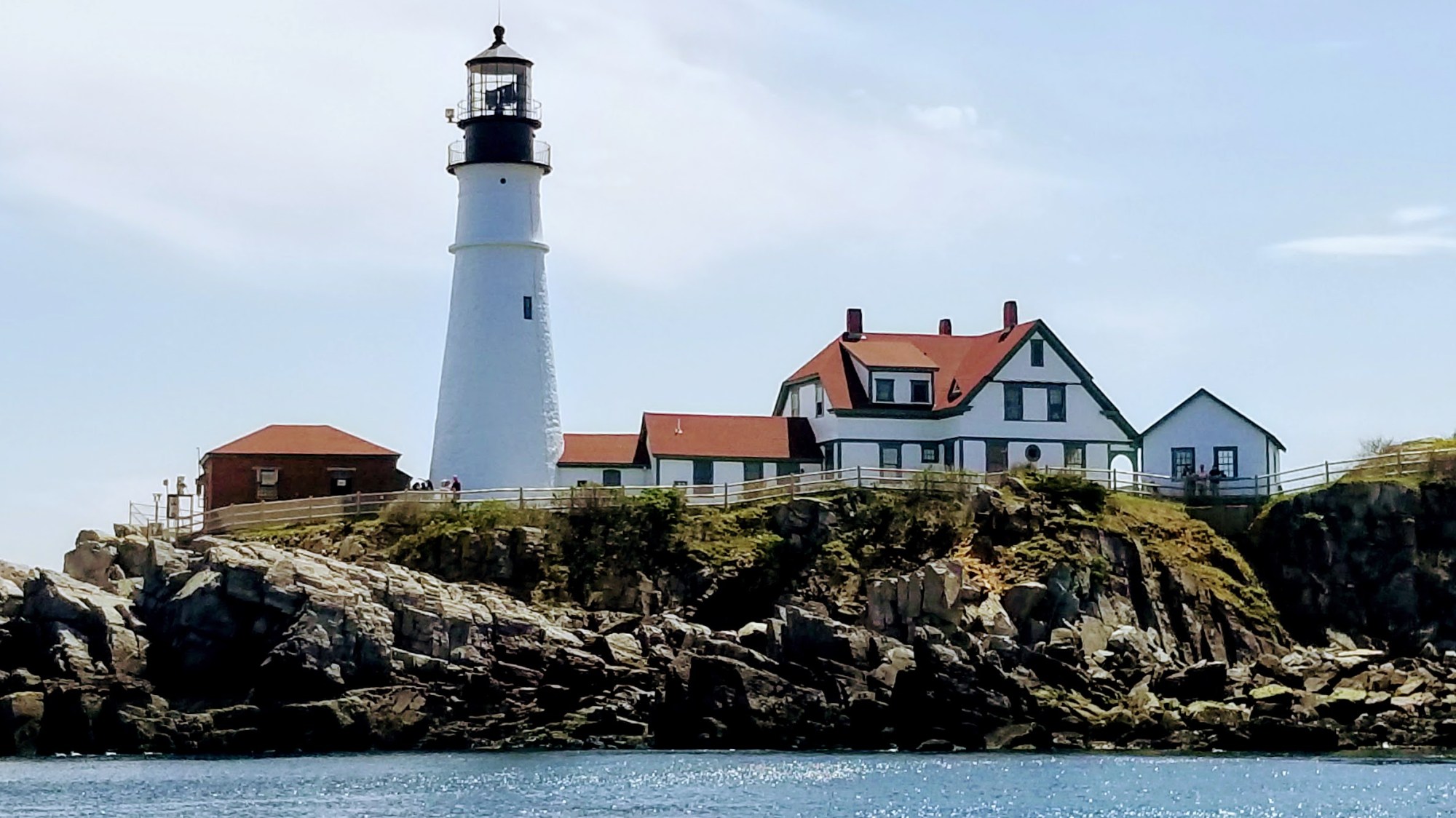 White lighthouse and house with red roofs on rocky coast under clear sky.