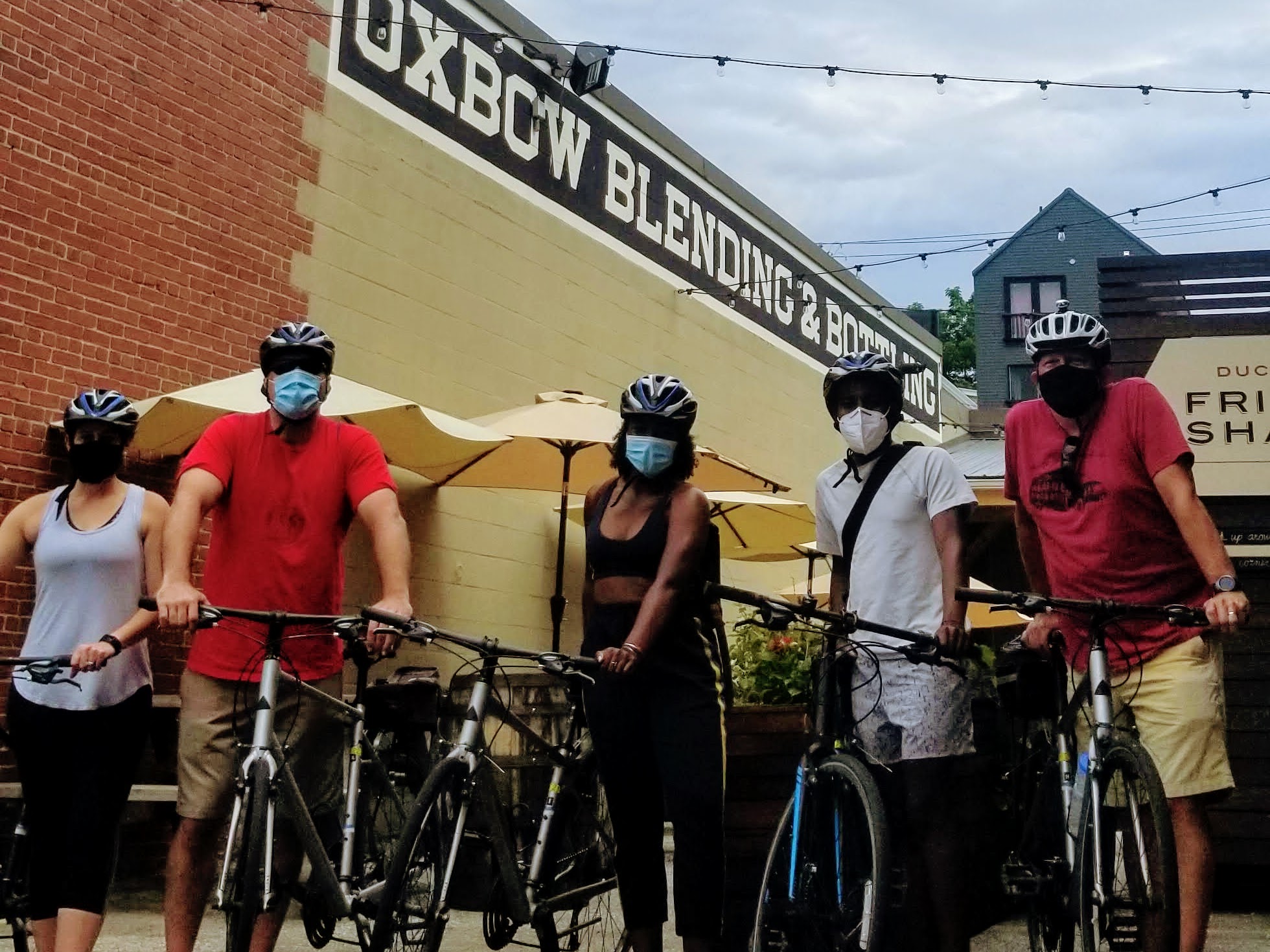 Five masked cyclists pose with bikes in front of Oxbow Blending & Bottling building.