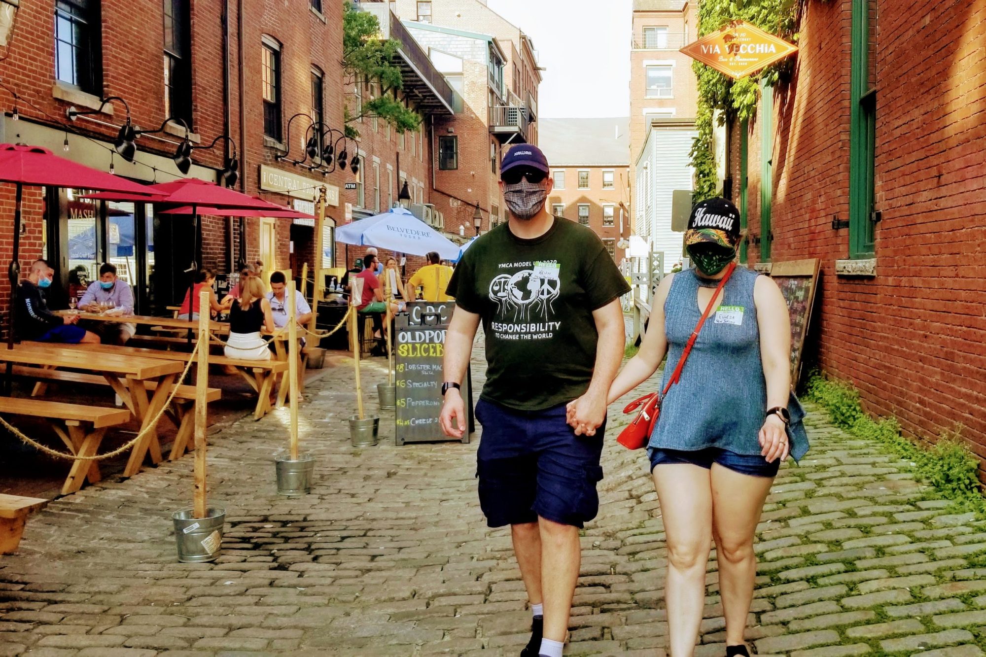 Masked couple holding hands, walking on a cobblestone street past outdoor diners.