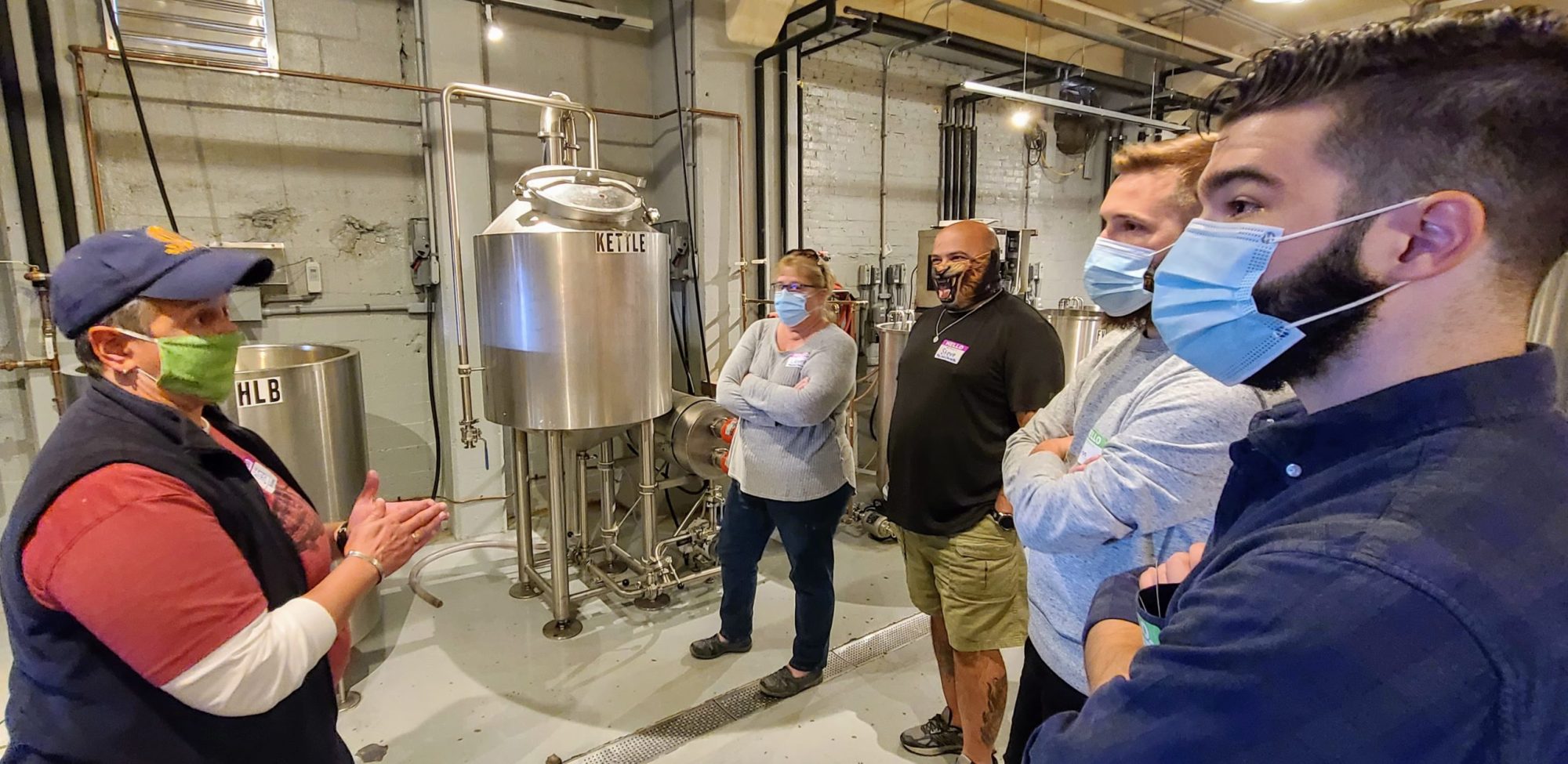 People in masks listen to a speaker in an industrial setting with large brewing equipment.