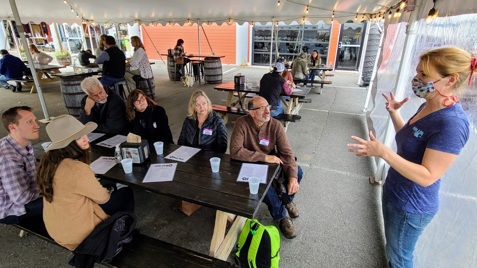 Group of people at picnic tables under a tent, listening to a woman speaking while wearing a mask.