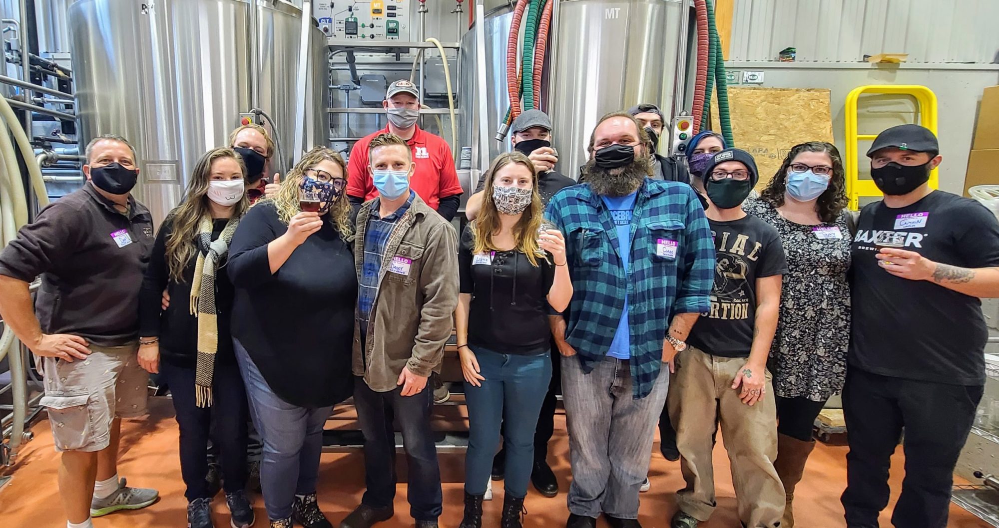 Group of people wearing masks standing in front of large brewery tanks, some holding drinks.