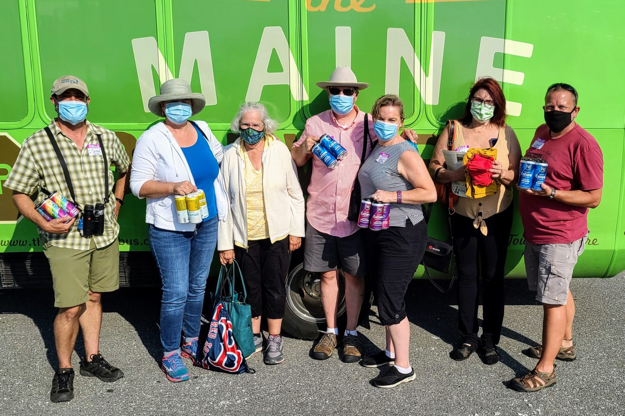 Seven people wearing masks stand in front of a green bus, holding canned beverages.