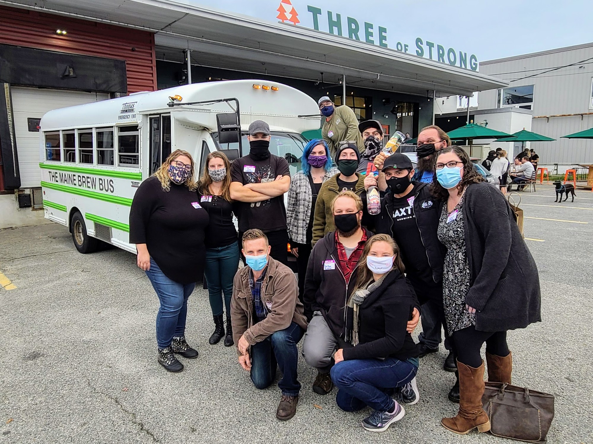 Group of people in masks pose in front of 'The Maine Brew Bus' and 'Three of Strong' building.