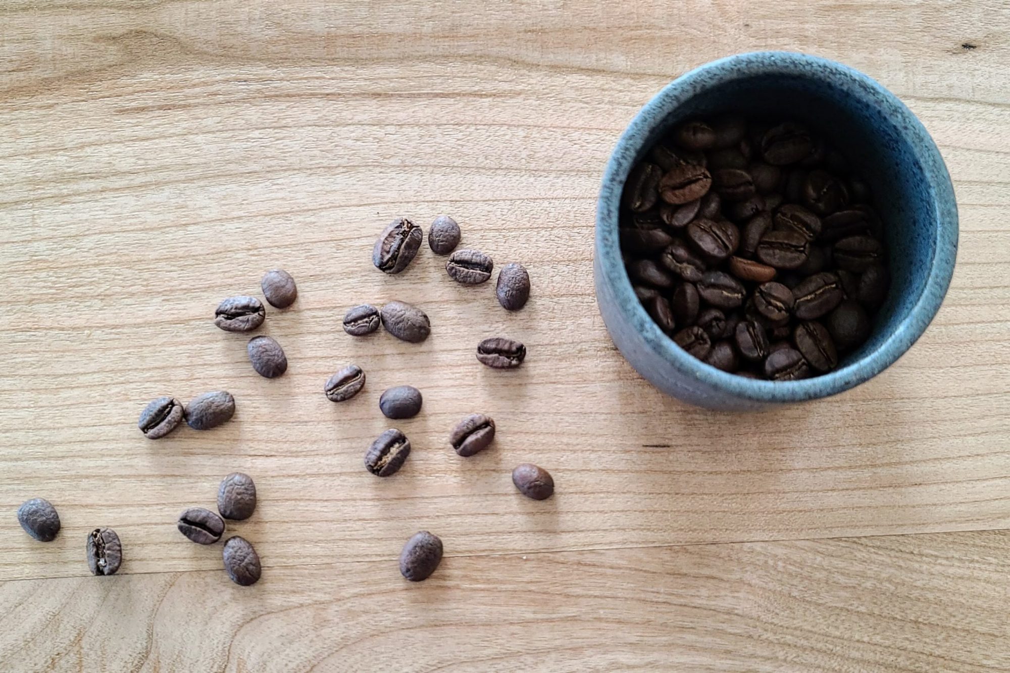 Coffee beans scattered on a wooden surface next to a blue cup filled with beans.