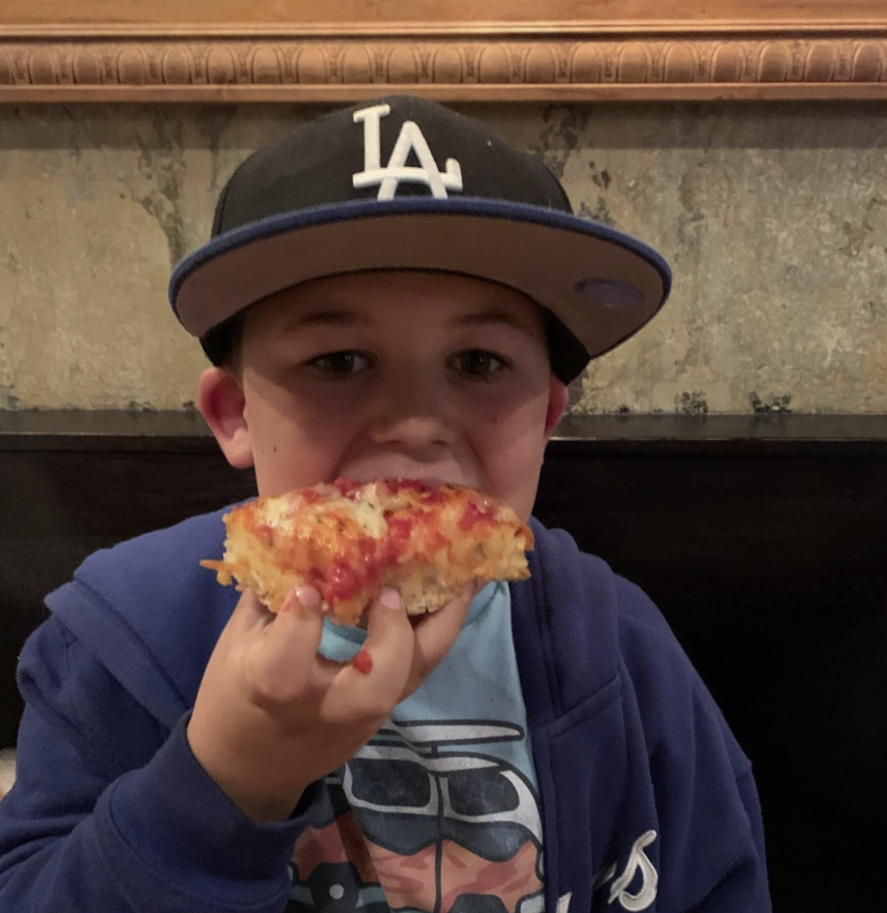 Child wearing a baseball cap eating a slice of pizza indoors.