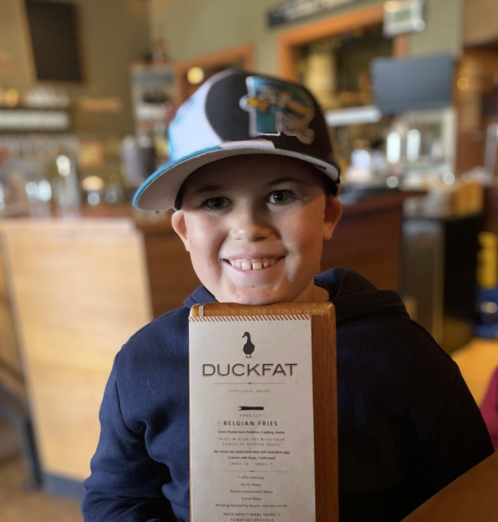 Child in a cap smiling, holding a Duckfat menu in a restaurant setting.