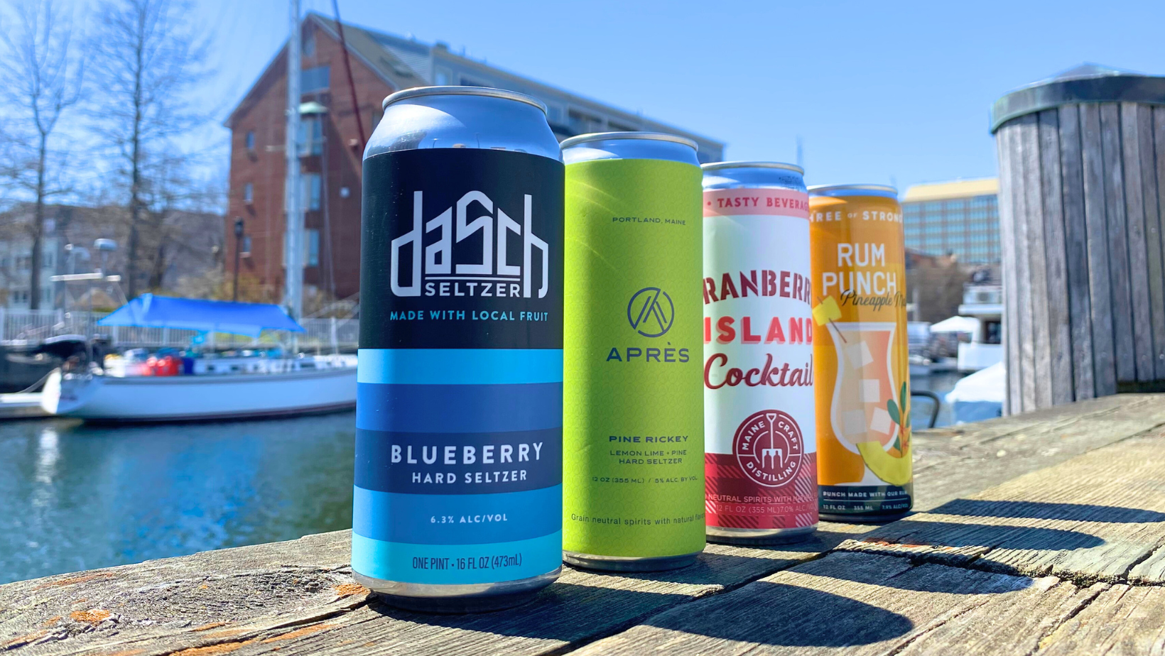 Four canned beverages on a wooden surface by water and buildings.