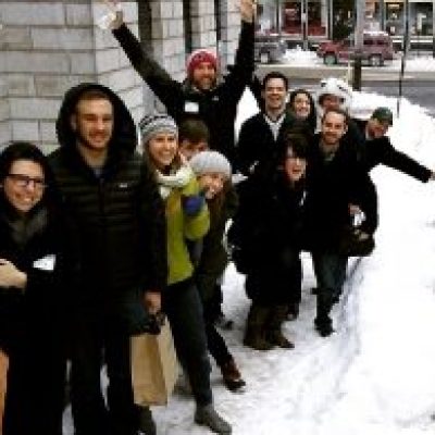 Group of people posing on a snowy sidewalk beside a building, waving and smiling.
