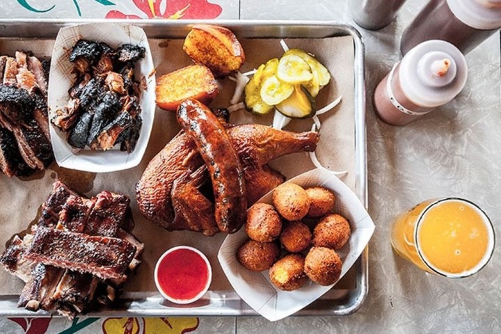 Tray of barbecue meats and sides with sauces and a drink on a table.