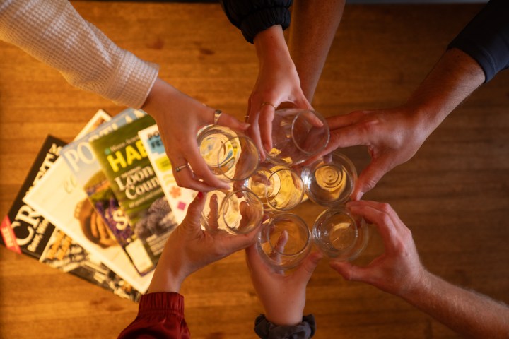 Six hands holding empty glasses in a toast above stacked magazines on a wooden surface.