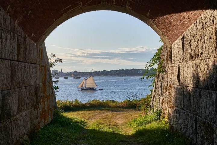 Sailboat on water viewed through stone archway with grassy foreground.