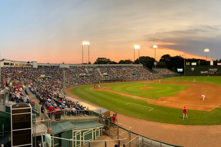 Baseball stadium with a game at sunset, large crowd in stands, field lights on.