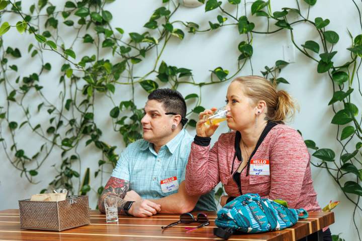 Two people sitting at a table in front of a leafy wall, one drinking from a glass.