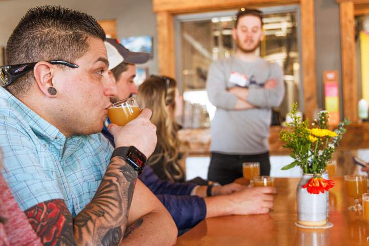 Person with tattoos drinks beer at a wooden table with a floral centerpiece, while others listen to a speaker.
