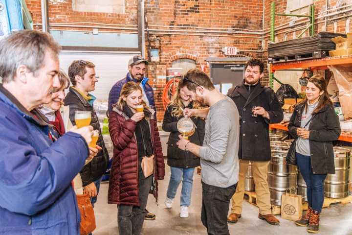 Group of people in a brewery tasting drinks, gathered around a person pouring beer.