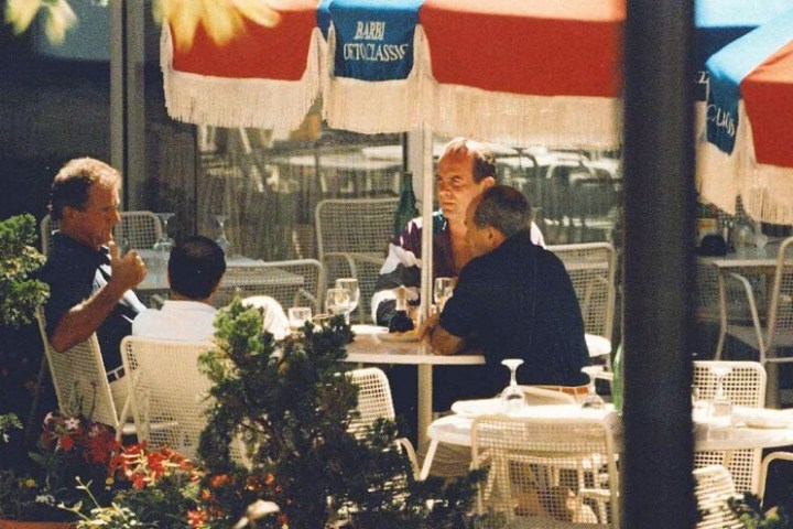 Four men sitting at an outdoor table under umbrellas, surrounded by plants.