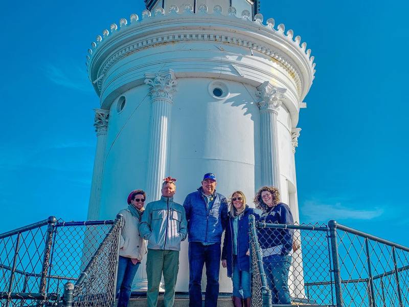 Five people stand in front of a white lighthouse with a blue sky.