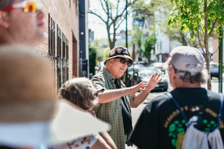 Group of people talking and gesturing on a sunny sidewalk, one wearing a hat and sunglasses.