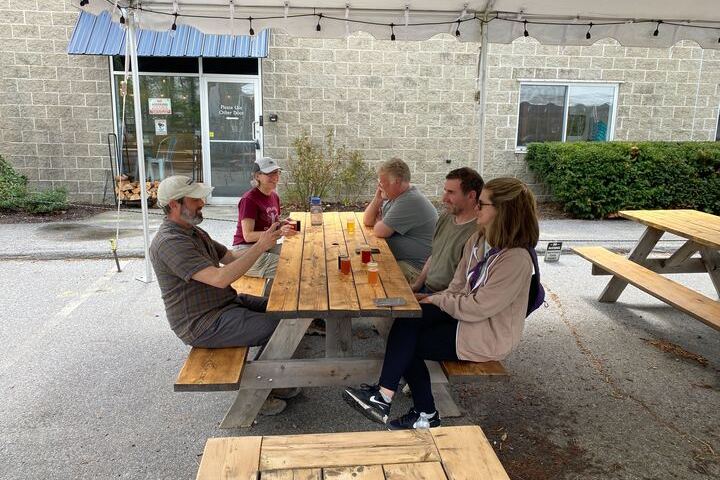 Five people sitting at an outdoor picnic table under a tent, drinking beverages.