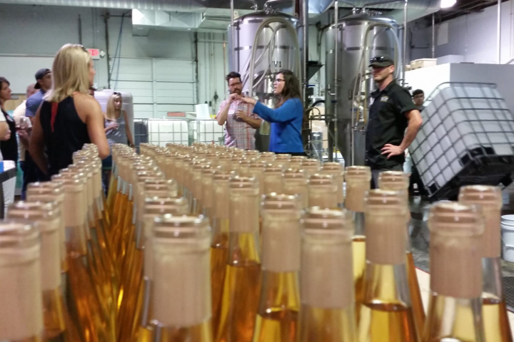 Tour group in a brewery, with rows of filled bottles in the foreground and brewing equipment in the background.