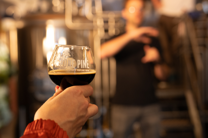 Person holding a glass of dark beer in a brewery setting, with blurred background.