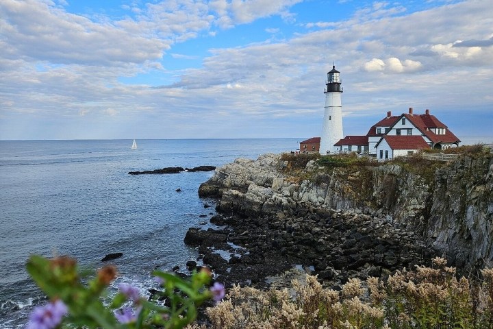 Lighthouse on rocky coast with red-roofed buildings and a sailboat on the ocean under a cloudy sky.