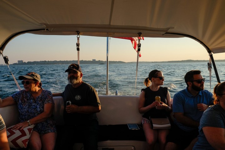 People sitting on a boat on a sunny day, with water and shoreline in the background.