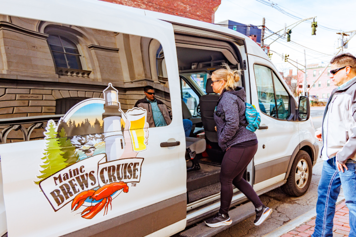 People boarding a white van with a 'Maine Brews Cruise' logo in an urban area.