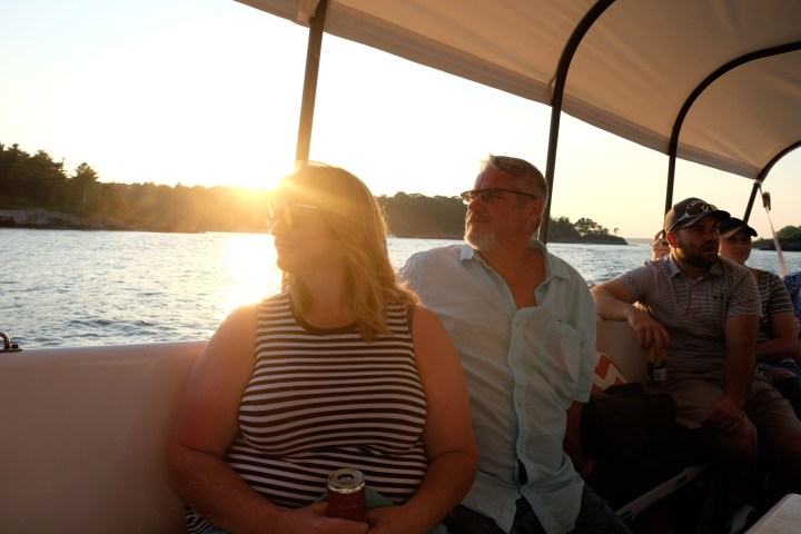 People on a boat at sunset, two looking outward, sun casting a warm glow.